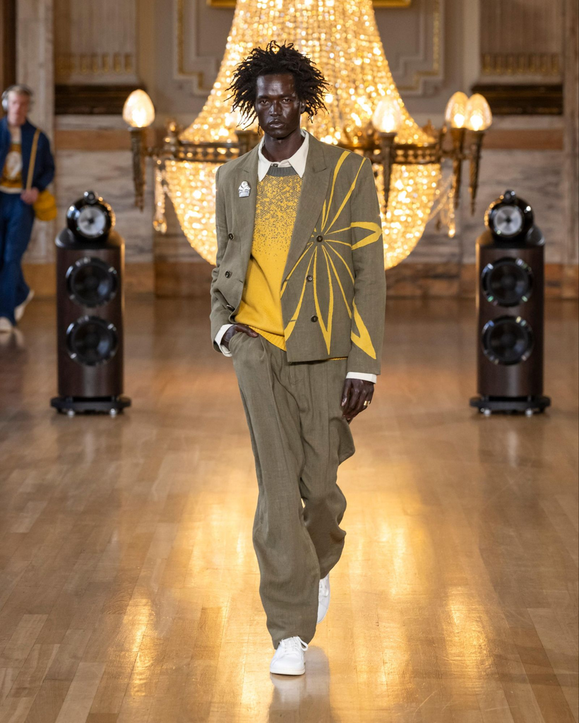 Dark-skinned male model with short locs, khaki suit jacket with yellow flower pattern on the right side, with matching khaki pants and white sneakers