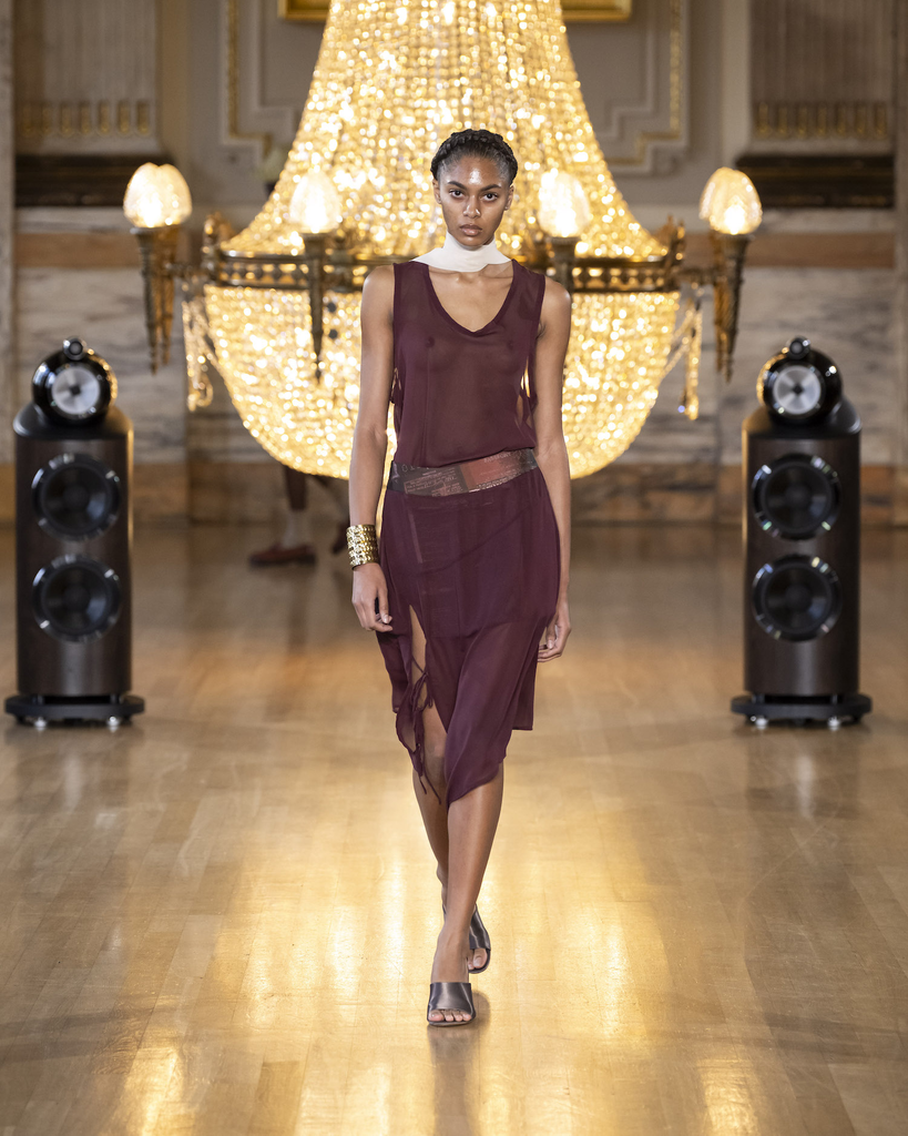Black female model in see-through burgundy dress, white silk scarf, large gold bangle on the right hand and open toe mules with thick heels